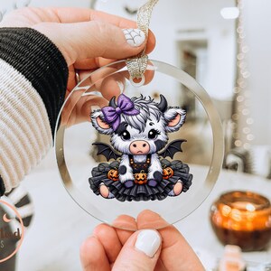 May include: A clear, round ornament featuring a cartoon cow with a purple bow, bat wings, and a black tutu. The cow is decorated with pumpkins, perfect for Halloween. The ornament is held by a person.