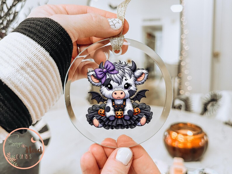 May include: A clear, round ornament featuring a cartoon cow with a purple bow, bat wings, and a black tutu. The cow is decorated with pumpkins, perfect for Halloween. The ornament is held by a person.