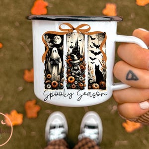 May include: White enamel mug with a black rim and handle, featuring a Halloween-themed design. The design includes a ghost, witch, black cat, and bats, with the words "Spooky Season" in a script font. The mug is held by a person.