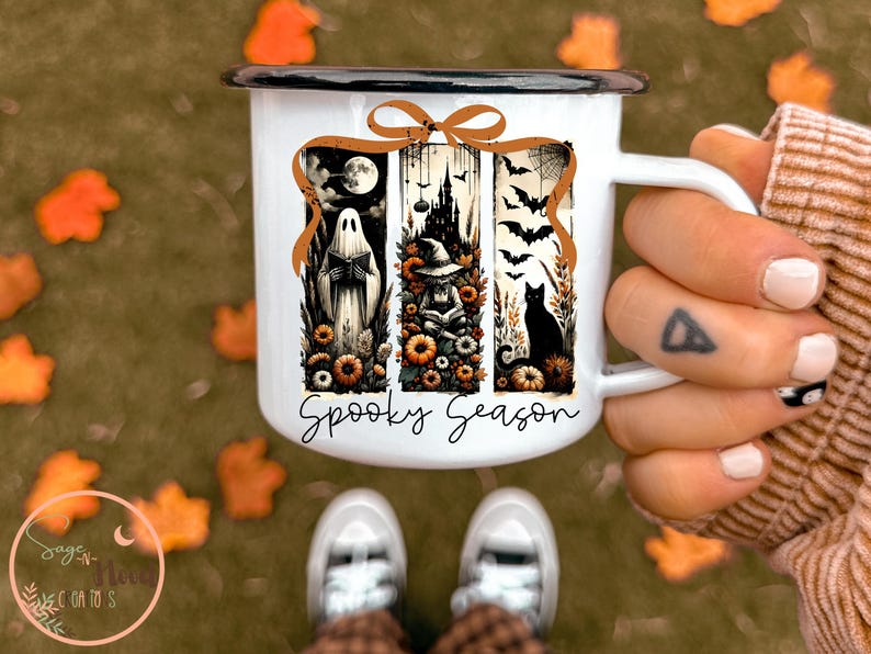 May include: White enamel mug with a black rim and handle, featuring a Halloween-themed design. The design includes a ghost, witch, black cat, and bats, with the words "Spooky Season" in a script font. The mug is held by a person.