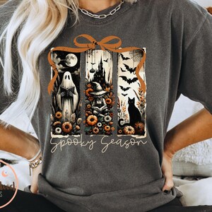 May include: Dark gray t-shirt with a Halloween-themed graphic. The design features a ghost, witch, and black cat within a framed design, with the words "Spooky Season" in a script font. The shirt has short sleeves.