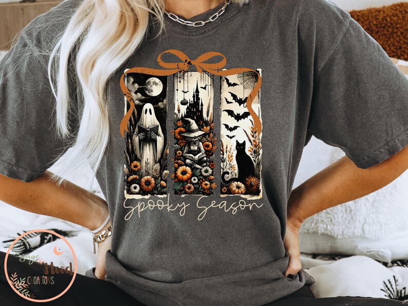 May include: Dark gray t-shirt with a Halloween-themed graphic. The design features a ghost, witch, and black cat within a framed design, with the words "Spooky Season" in a script font. The shirt has short sleeves.