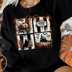 May include: Black sweatshirt featuring a four-panel Halloween design. The panels include a jack-o'-lantern, a ghost, a haunted house, and the words "Spooky Season". The design is framed with a brown border.