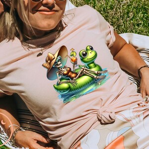 Może przedstawiać: R&oacute;żowy t-shirt z grafiką szkieletu w kapeluszu przeciwsłonecznym, jadącego na nadmuchiwanej zielonej żabie z koktajlem. Żaba ma na sobie zielony kapelusz.