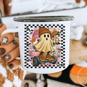May include: White enamel mug with a black rim. The mug features a cartoon ghost on a hoverboard, giving a thumbs up. The design includes a checkerboard background, bats, pumpkins, and the text "WE WEAK".