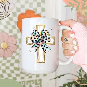 May include: White ceramic coffee mug with a gold cross and a colorful polka dot bow. The bow is pink, blue, and yellow with black polka dots.