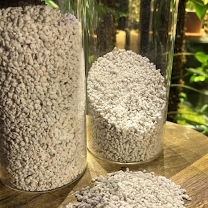 Pode incluir: Dois frascos de vidro cheios de material branco pequeno semelhante a pedras. Um monte do mesmo material está sobre uma superfície de madeira.