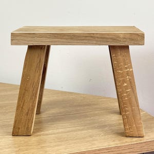 May include: A small, light brown wooden stool. It has a rectangular top and four angled legs. The stool is made of natural wood and has visible grain patterns. It is designed for use as a footstool or plant stand, with dimensions suitable for small spaces.