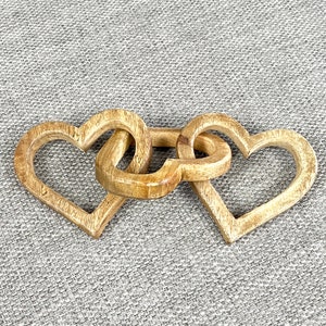 May include: Three wooden hearts interlocked together, forming a decorative chain. The hearts are a light brown colour and have a smooth, polished finish.