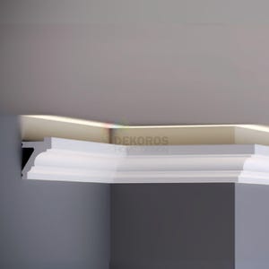 May include: White crown molding installed on a wall and ceiling. The molding has a decorative design and is positioned at a corner. The text "DEKOROS HOME DESIGN" is visible on the wall.