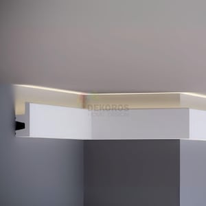 Peut inclure: Un design de plafond blanc moderne avec éclairage encastré. L'image montre un coin de pièce avec une structure de plafond en couches. Le texte "DEKOROS HOME DESIGN" est visible. Le design intègre des éléments blancs et gris.