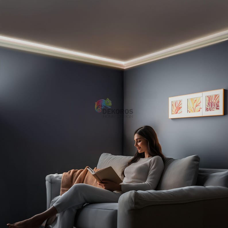 May include: A woman reading a book while sitting on a gray sofa in a room with a decorative ceiling light. The room has dark gray walls and a framed artwork with three panels.