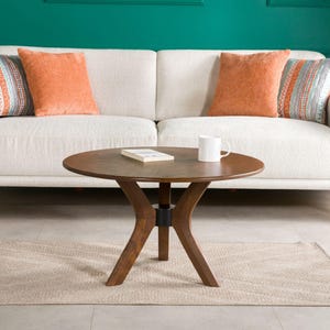 Può includere: Un salotto con un divano bianco adornato da cuscini arancioni e fantasia. Un tavolino rotondo in legno espone un libro e una tazza bianca. Un tappeto beige e un oggetto decorativo arancione completano l'ambiente.