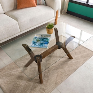 May include: A rectangular glass coffee table with a dark wood base. The base has a unique design with angled legs. A magazine and small plant sit on the table. The table is on a beige rug in a living room setting.
