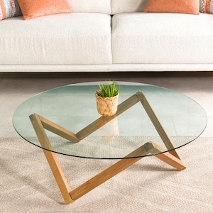 May include: A round glass coffee table with a wooden base. The base is made of four pieces of wood that are arranged in a criss-cross pattern. The table is sitting on a light brown rug.