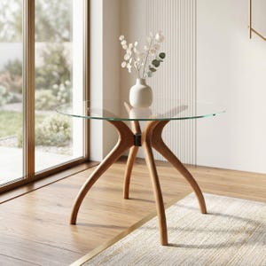 May include: A round glass-top table with a light brown wooden base featuring four curved legs that meet at a central black connector. A white ceramic vase containing dried stems with small round leaves sits on the center of the glass surface.