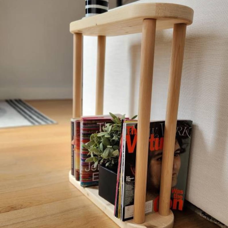 May include: A wooden side table with two shelves. The top shelf is empty and the bottom shelf holds a potted plant and several magazines. The magazines have titles like "The New Yorker" and "Virtue".