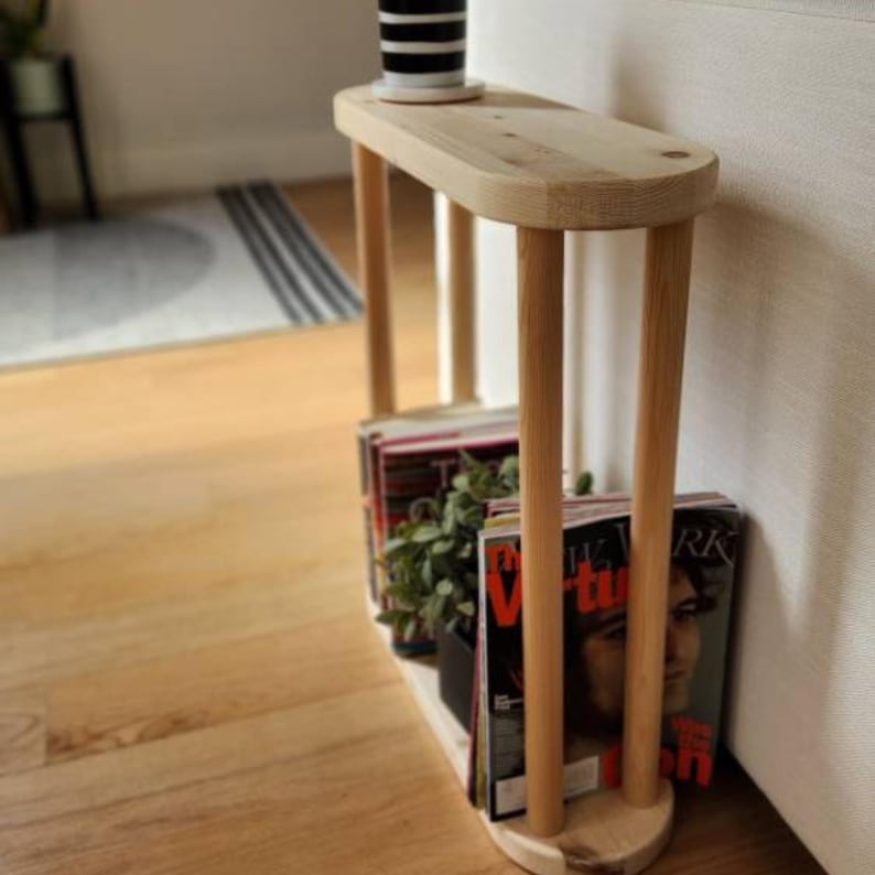 May include: A wooden side table with a rounded top and four legs. The table is made of natural wood and has a rustic look. It is holding a stack of magazines and a small potted plant.