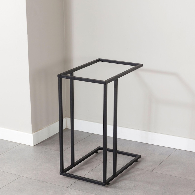 May include: A black metal side table with a rectangular top and a geometric frame. The table has a C-shaped design, with a base that is 15 inches long and 10 inches wide. The table is 24 inches tall.