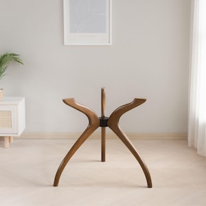 Könnte beinhalten: Ein Tischfuß aus Holz im modernen Mid-Century-Design. Der Fuß hat drei gebogene Beine und eine zentrale Stützsäule, alles in einem warmen Braunton. Ein schwarzer Ring verbindet die Beine mit der Mittelsäule.