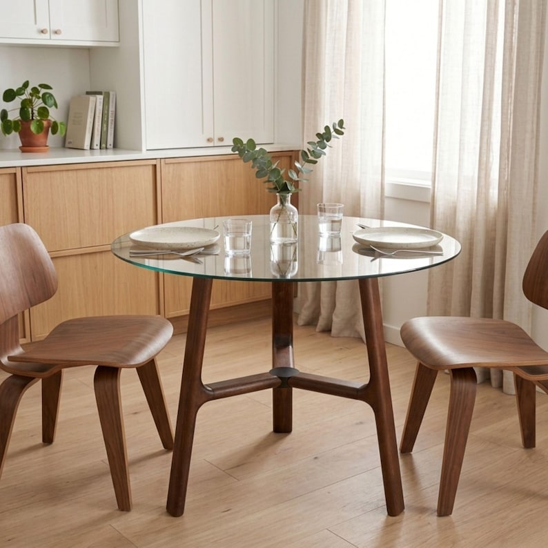 May include: A round dining table with a clear glass top and a dark brown wooden base with three legs, accompanied by two matching curved wooden chairs. A clear glass vase with green eucalyptus stems sits in the center of the table.