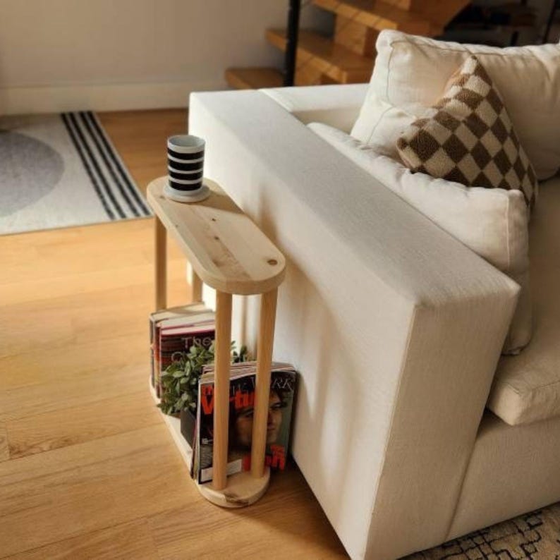 May include: A light wood side table with two shelves, one holding books and magazines, the other holding a plant and a mug. The table is next to a white couch.