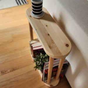 May include: A wooden side table with a rounded top and a shelf underneath. The table is made of natural wood and has a light brown finish. The table is next to a white couch and has a white mug and a stack of books on it.