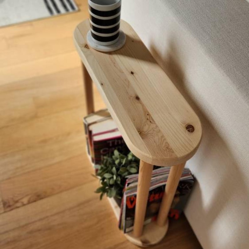 May include: A wooden side table with a rounded top and a shelf underneath. The table is made of natural wood and has a light brown finish. The table is next to a white couch and has a white mug and a stack of books on it.