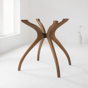 Può includere: Una base per tavolo in legno marrone caratterizzata da quattro gambe curve e affusolate che si estendono da un perno centrale. La struttura è realizzata in legno liscio e levigato con venature visibili, progettata per sostenere un piano d'appoggio.