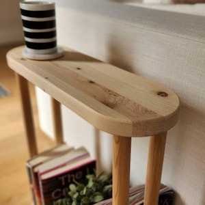 May include: A wooden side table with a rounded oval top and two legs. The table is made of light wood and has a natural finish. The table is sitting next to a couch and has a stack of books and a plant on the bottom shelf.