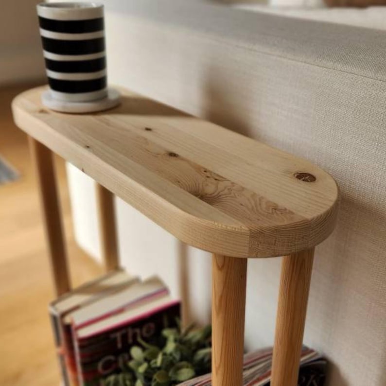 May include: A wooden side table with a rounded oval top and two legs. The table is made of light wood and has a natural finish. The table is sitting next to a couch and has a stack of books and a plant on the bottom shelf.