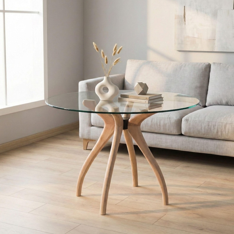 May include: A round glass-top table with a light wood base. The table has a clear glass surface and three curved wooden legs. A vase with dried flowers and a stack of books are on the table. The table is near a window and a gray sofa.