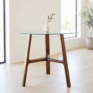 May include: A round glass-topped table with a dark brown wooden base featuring three legs connected by a central hub. A small white ceramic vase containing a single green leafy branch sits on the center of the clear glass tabletop surface.