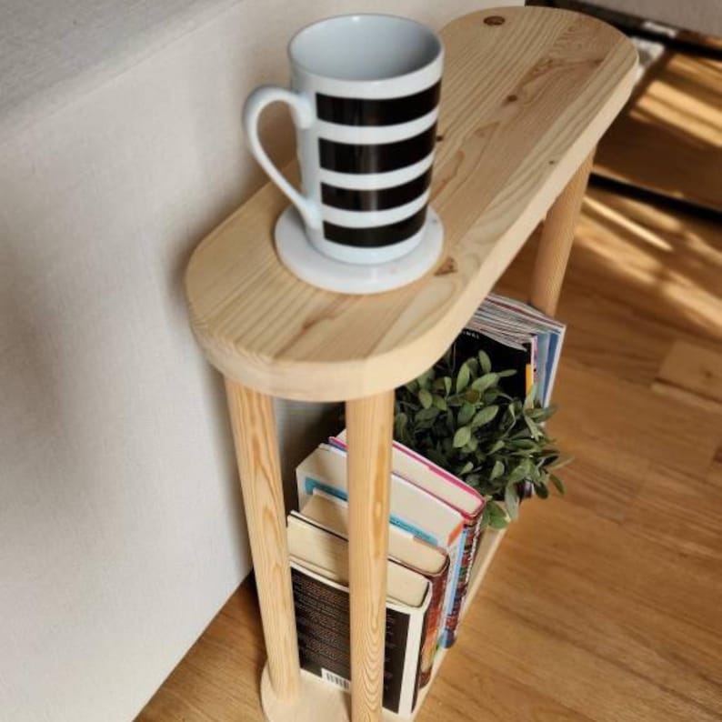 May include: A wooden side table with a rounded top and a shelf underneath. The table is made of natural wood and has a white mug with black stripes on top. The shelf is filled with books and a small green plant.