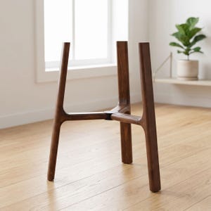 May include: A dark brown wooden plant stand featuring three vertical legs connected by a central horizontal support structure. The stand has a smooth, polished finish and a sturdy, geometric design intended to hold a container or pot securely.
