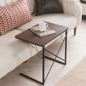 Op de afbeelding: Een donkerbruine bijzettafel met een rechthoekig blad en een zwart metalen frame. Een witte mok en een tijdschrift liggen op de tafel. De tafel staat naast een beige bank met decoratieve kussens.