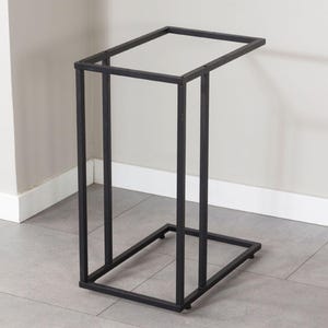 May include: A black metal side table with a rectangular top and a C-shaped design. The table has a minimalist style, with a sturdy frame and a sleek, modern aesthetic. The table is designed to slide under a sofa or chair.