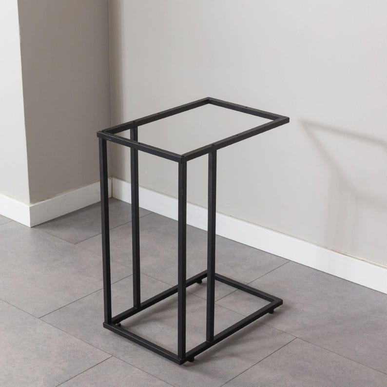 May include: A black metal side table with a rectangular top and a C-shaped base. The table is made of square metal tubing and is designed to slide under a sofa or chair. The table measures approximately 24 inches high, 12 inches wide, and 20 inches deep.