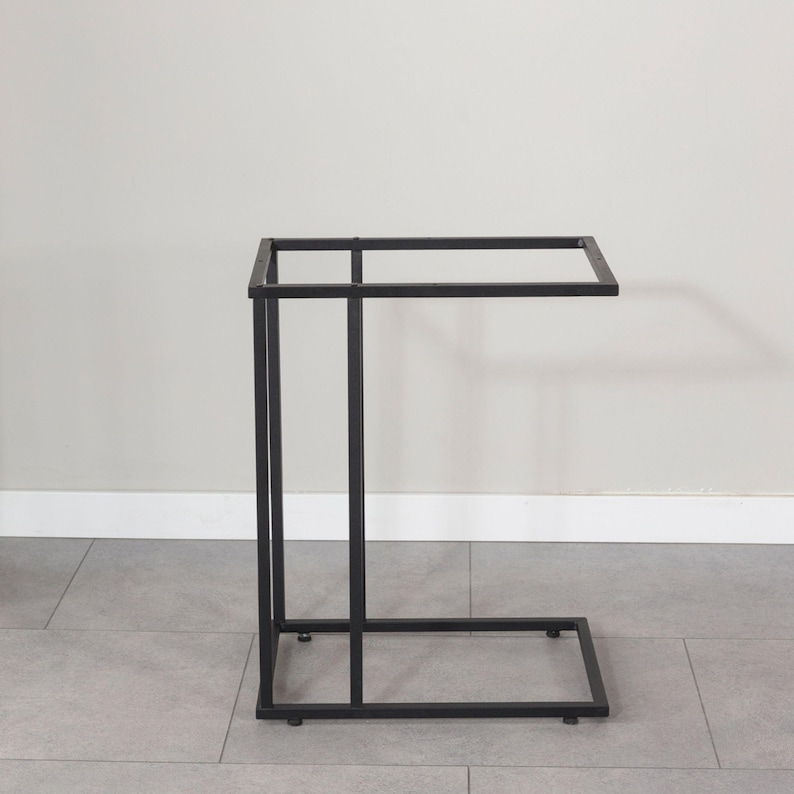 May include: A black metal side table with a rectangular top and a C-shaped base. The table is approximately 24 inches long, 12 inches wide, and 24 inches high. The design is minimalist and modern, suitable for various decor styles.
