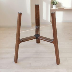 May include: A dark brown wooden plant stand featuring three vertical legs connected by a central hub. The structure is designed to hold a container, with each leg extending upward from a reinforced circular base joint in the center.