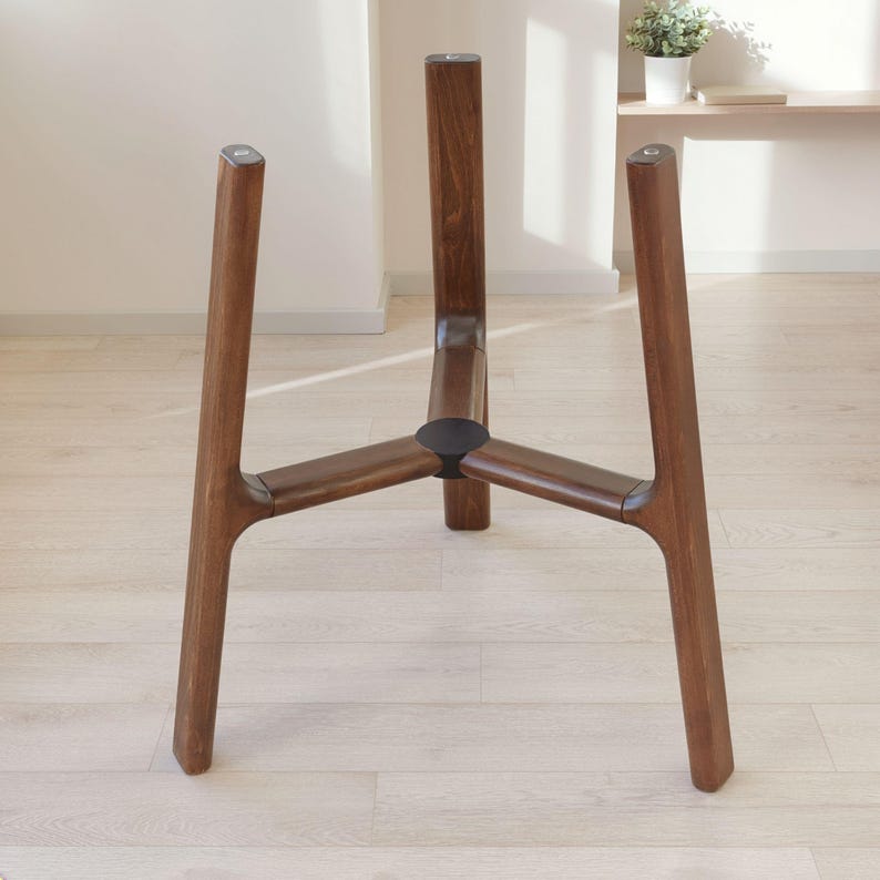 May include: A dark brown wooden plant stand featuring three vertical legs connected by a central hub. The structure is designed to hold a container, with each leg extending upward from a reinforced circular base joint in the center.