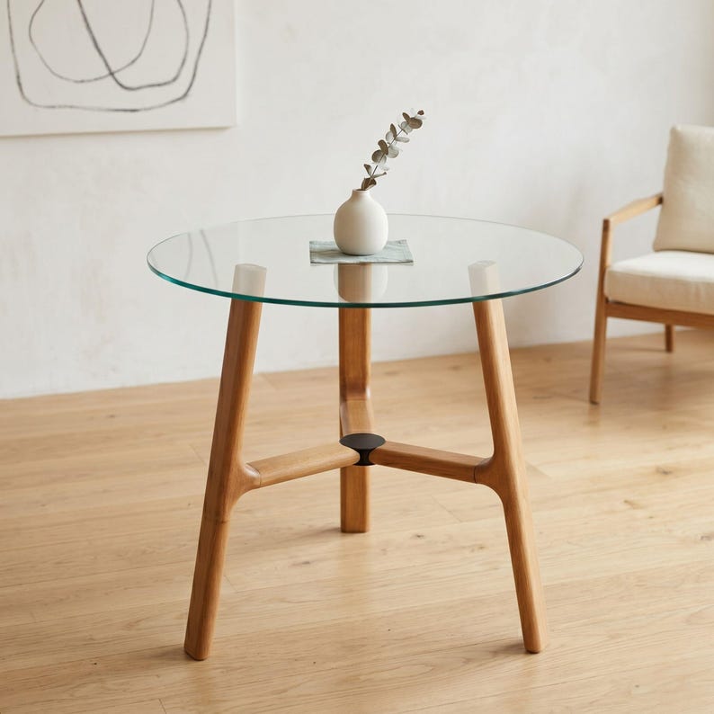 May include: A round glass-topped table with a light-toned wooden base featuring three angled legs connected by a central hub. A small white ceramic vase containing a single sprig of eucalyptus sits centered on the clear glass surface.