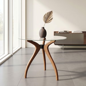 May include: A round glass-top table with a light brown wooden base featuring three curved legs that meet at a central black cylindrical hub. A dark brown ceramic vase sits on the table, holding a single dried, fan-shaped palm leaf.