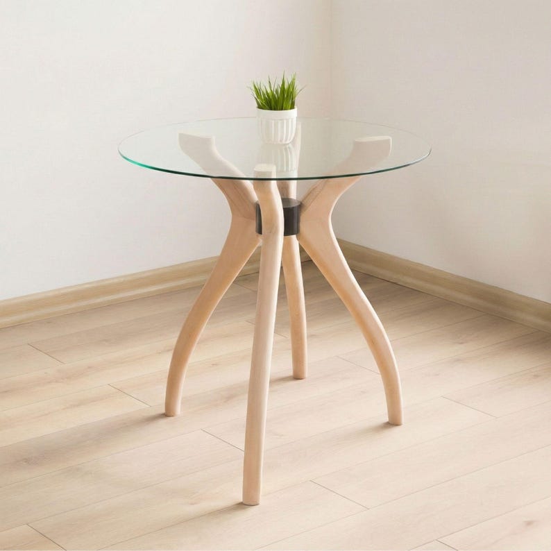 May include: A modern side table with a clear glass top and light wood legs. The legs are curved and splay outwards, providing a unique design. A small white pot with green grass sits on the table. The table is approximately 24 inches in diameter.