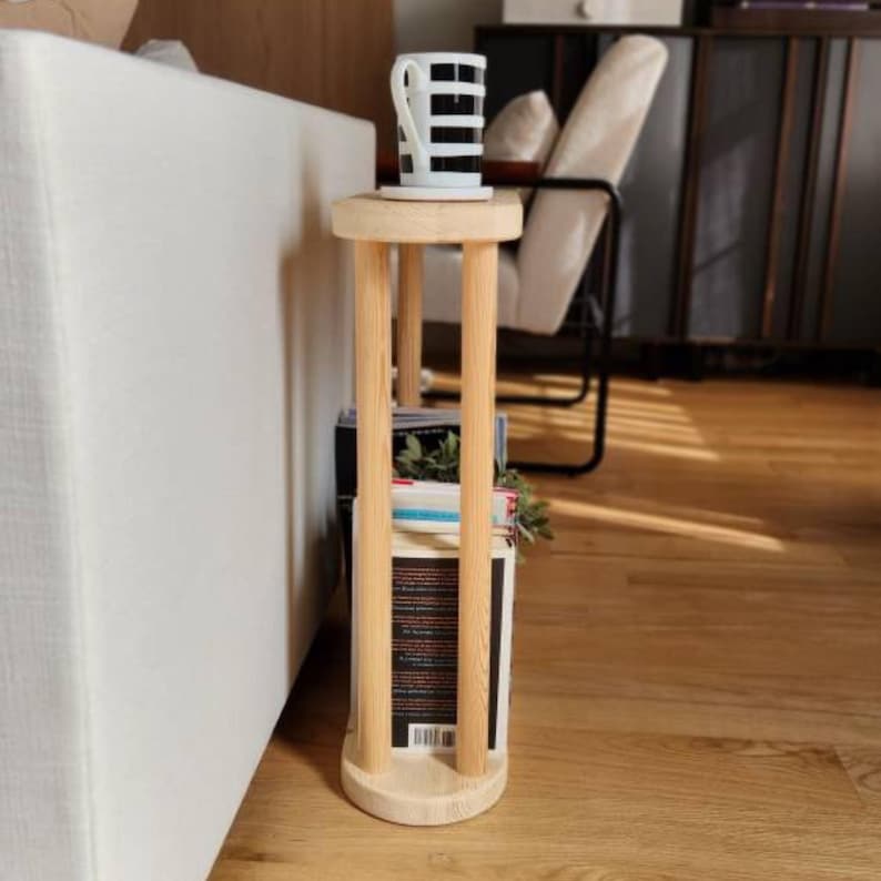May include: A wooden side table with a natural finish. The table has a round top and four vertical supports. The table is holding a stack of books and a white mug with black stripes.