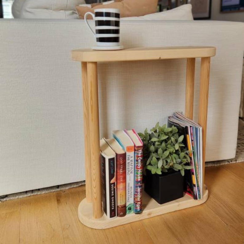 May include: A light wood end table with two shelves. The top shelf has a white mug with black stripes. The bottom shelf has a potted plant, a stack of books, and a stack of magazines. The books have titles like "The Candy House" and "Seeing Ghosts".