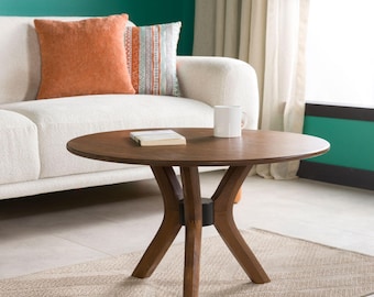 May include: A round wooden coffee table with a smooth brown finish and four curved legs meeting at a central base. A white ceramic mug and a closed rectangular book are placed on the tabletop surface, which is positioned in front of a sofa.