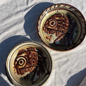 May include: Two small, round ceramic bowls with a fish design. The fish are depicted in shades of brown and tan, with a large eye and detailed scales. The bowls have a cream-colored background and a brown rim.