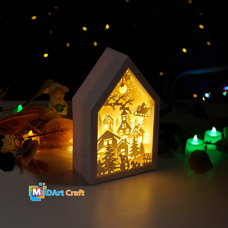 May include: A white wooden house-shaped light-up decoration with a cut-out design of a winter village scene, including houses, trees, a snowman, and Santa Claus in his sleigh. The light shines through the cut-outs, creating a warm glow.