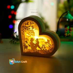 May include: A white heart-shaped wooden lamp with a cutout design of a mother and child sitting in a park with a teddy bear. The lamp is illuminated from within, casting a warm glow.